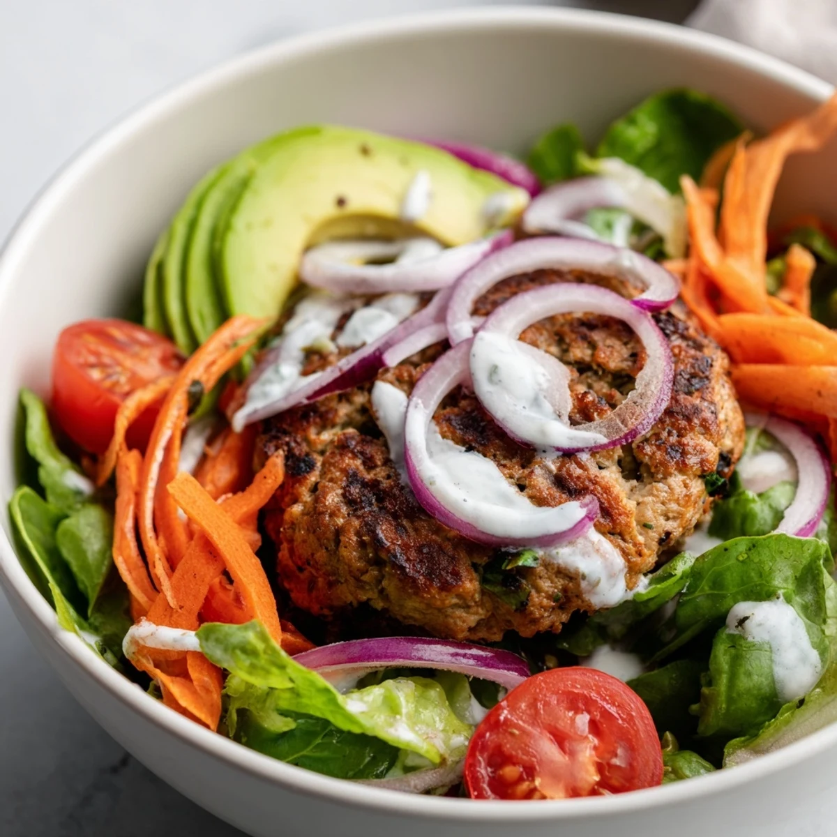 Juicy turkey burger patties rest on a vibrant salad bowl filled with crisp greens, cherry tomatoes, cucumber, and creamy avocado, topped with tangy yogurt dressing.  