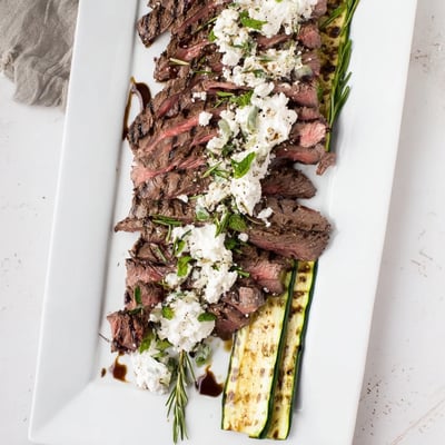 Sliced tender balsamic flank steak served over creamy whipped feta with charred zucchini on a platter.